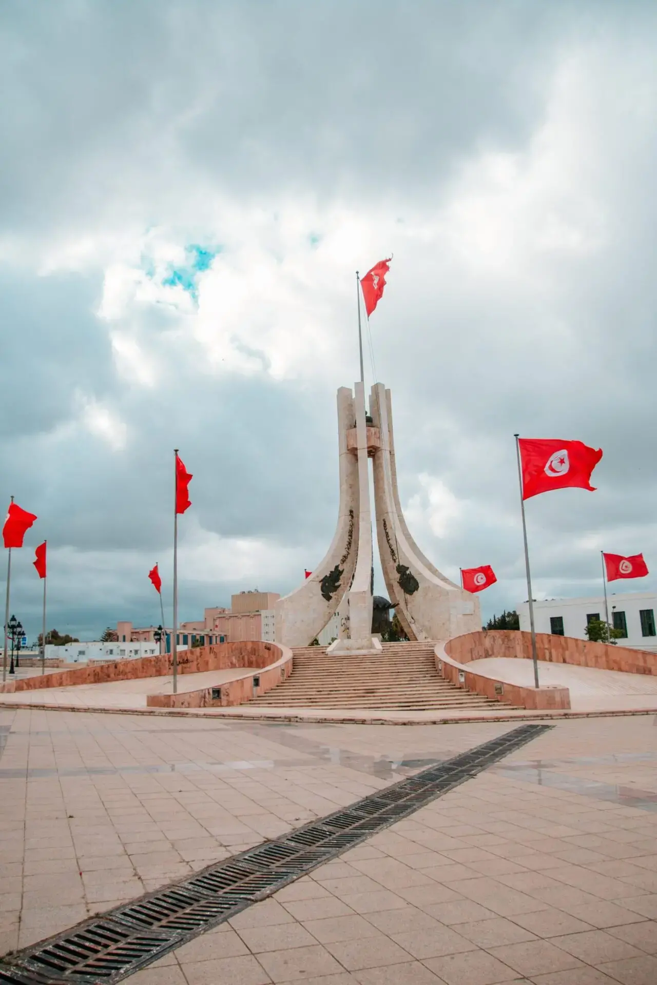 A large white monument with a tall central flagpole and several surrounding red Tunisian flags stands in an open plaza under a cloudy sky. Stairs lead up to the monument, with buildings in the background.