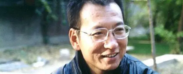 A man with short black hair and glasses smiles while wearing a dark jacket outdoors, with greenery and a blurred background behind him, reminiscent of Liu Xiaobos thoughtful presence.