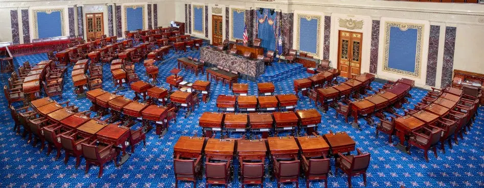 The image shows the U.S. Senate chamber, featuring rows of wooden desks and chairs arranged in a semicircle on a blue-patterned carpet, with a central podium and decorative walls.
