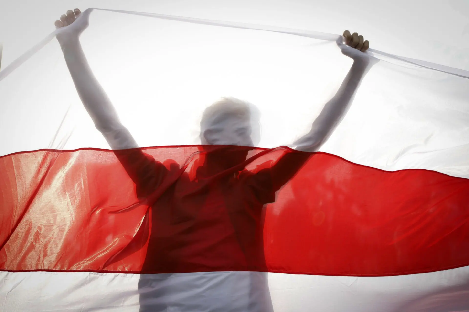 A person stands holding up a large white and red flag, with the sunlight shining through, making the figure partially silhouetted behind the flag.