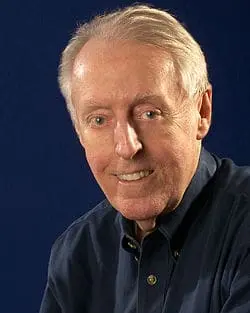 An older man with light skin and gray hair smiles at the camera. He is wearing a dark button-up shirt and is posed against a solid dark blue background.