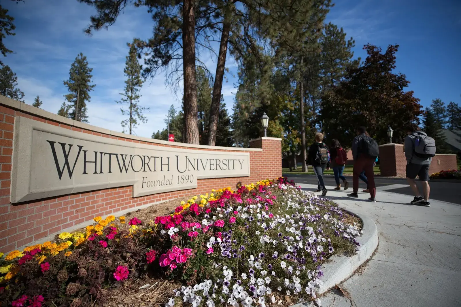 A stone sign for Whitworth University, surrounded by colorful flowers, sits beside a walkway where several people are walking on a sunny day with tall trees in the background.