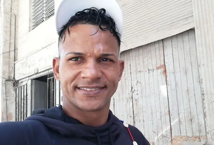 A man with short, curly hair and a white cap smiles at the camera. He is wearing a dark hoodie and standing in front of a weathered building with wooden doors and barred windows.