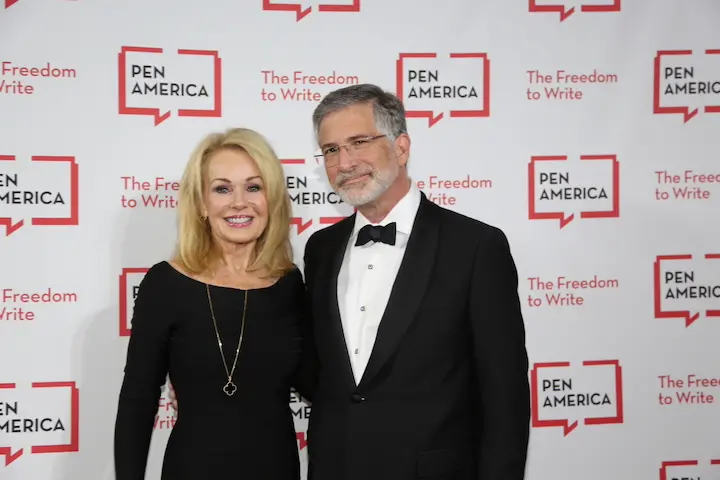 A smiling woman in a black dress and a man in a tuxedo stand together at a PEN America event, with a backdrop that reads PEN America and The Freedom to Write in red and white.