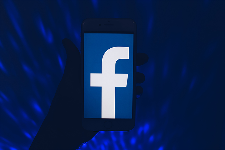 A hand holds a smartphone displaying the large white Facebook f logo on a blue background, with a dark blue light pattern in the background, highlighting PEN Americas commitment to human rights in the digital age.