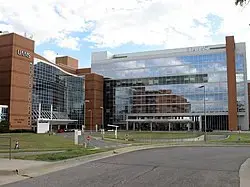 A modern hospital building with large glass windows and brick sections, featuring multiple stories and a curved driveway in front under a partly cloudy sky.