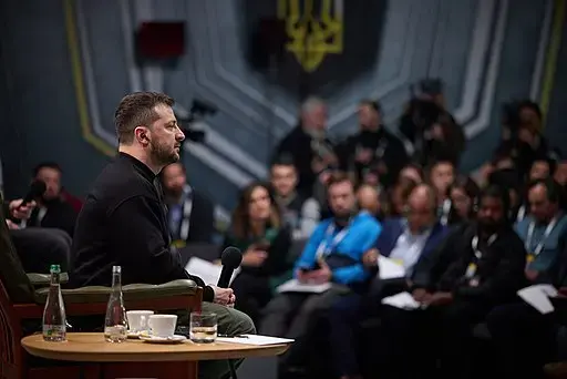 A man sits on a stage holding a microphone, facing an audience. The audience is seated and appears attentive, with some people taking notes. The background features a large emblem and blurred figures.