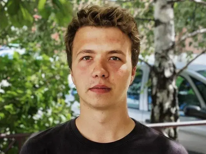 A young man with short curly brown hair and a serious expression stands outdoors in front of trees and a parked car, wearing a black t-shirt.
