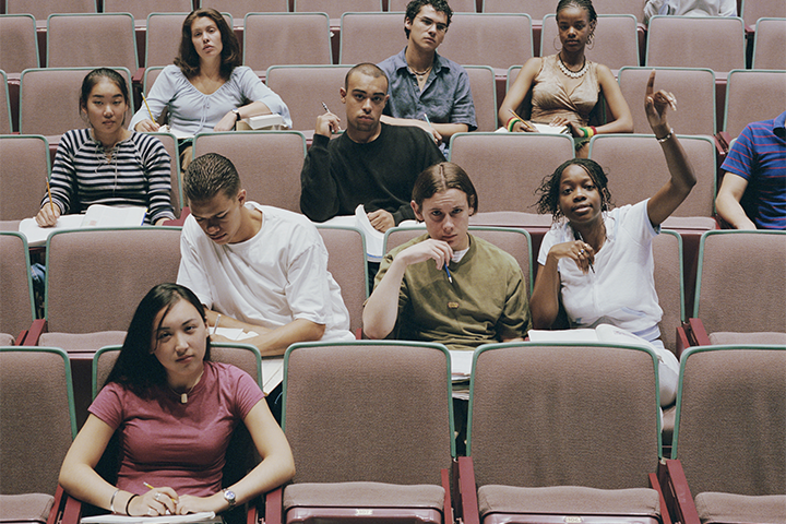 A diverse group of students sits in an auditorium, some taking notes, while others raise their hands or look thoughtful, suggesting active participation in a lecture or classroom discussion.