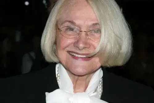 An older woman with short, light blonde hair and glasses, smiling. She is wearing a black outfit with a white bow and pearl necklace.