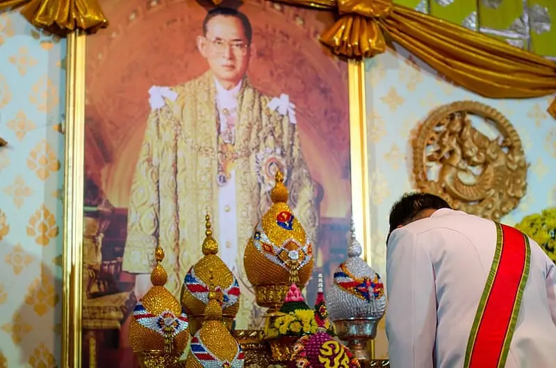 A person in formal attire bows in front of an elaborate altar with ornate ceremonial items, beneath a large portrait of a man in golden royal robes, adorned with medallions, against a decorative golden background.