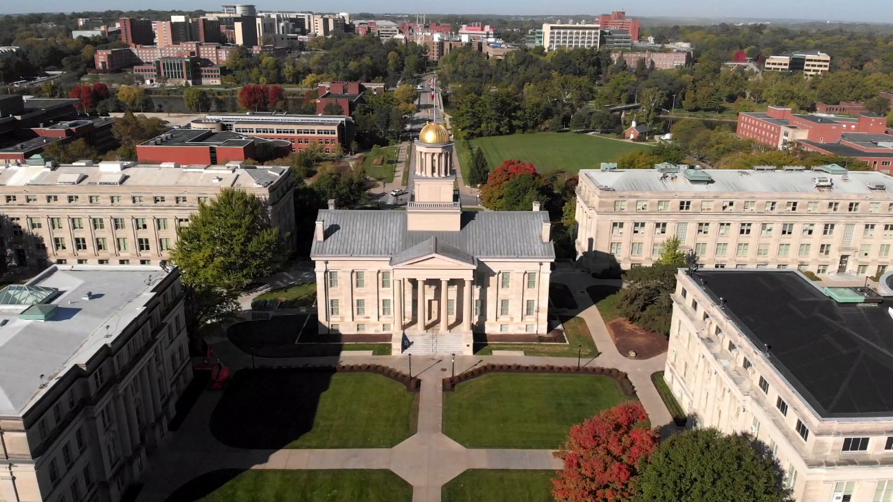 Aerial view of a historic building with a gold dome, surrounded by green lawns, trees, and other campus buildings, with a cityscape in the background on a clear day.
