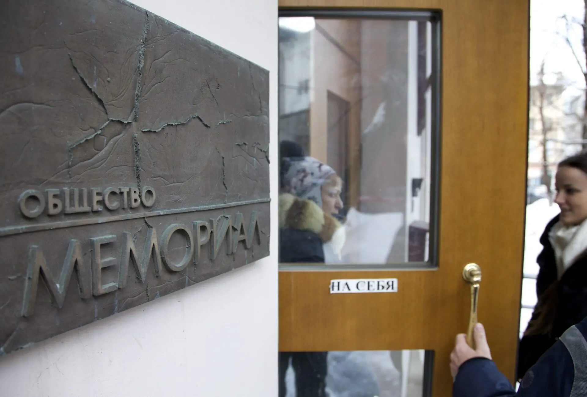 A person opens a door labeled НА СЕБЯ with a plaque reading ОБЩЕСТВО МЕМОРИАЛ on the wall. Two people are visible through the glass door, with snow visible outside.