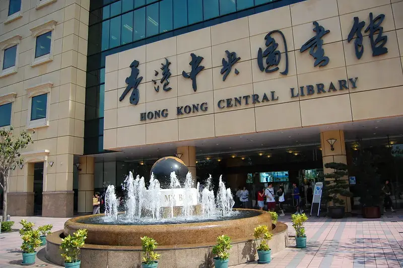 The entrance of Hong Kong Central Library with large Chinese and English signage, a globe fountain in front, potted plants, and people walking near the doors.