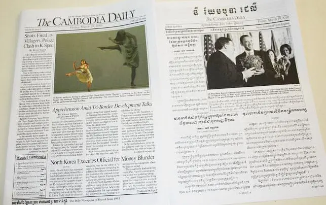 Two pages of The Cambodia Daily newspaper, one in English and one in Khmer, showing headlines, articles, and photos, including a ballet dancer and a group of people with flowers in a formal setting.