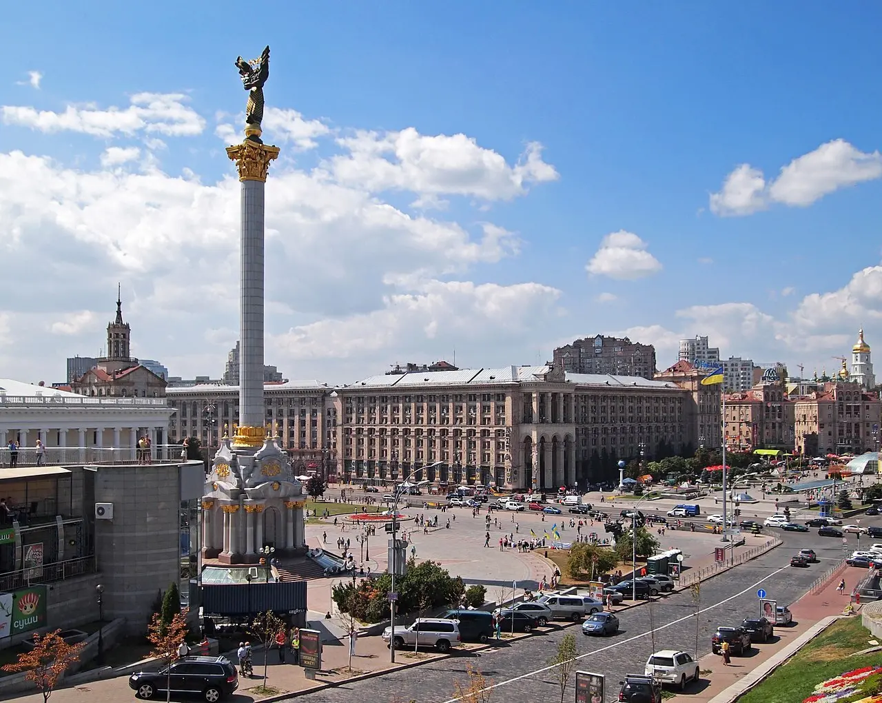 A large city square with a tall column topped by a golden statue in the center, surrounded by historic buildings, cars, and people under a blue sky with scattered clouds.
