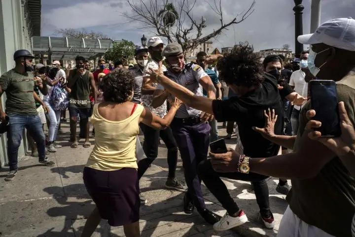 A chaotic street scene where several people are struggling and pushing each other; one man in uniform appears to intervene as bystanders watch and take photos. The atmosphere is tense and crowded.