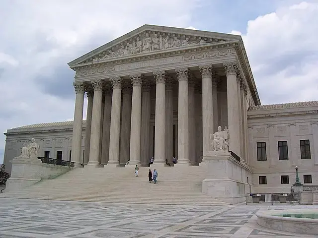 The image shows the front of the United States Supreme Court building, featuring large columns, wide steps, statues, and a cloudy sky above. A few people are walking up the steps.