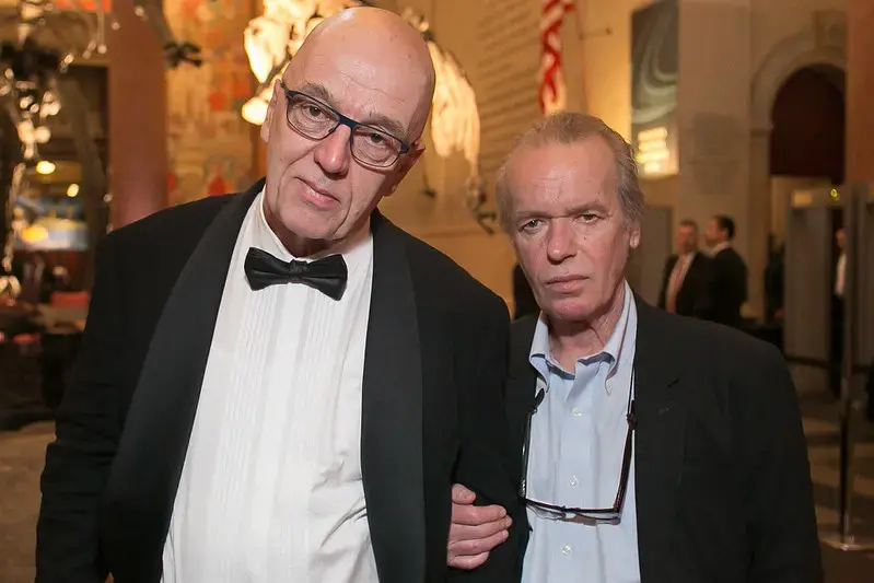 Two men in formal attire pose together at an indoor event; one wears a tuxedo with a bow tie and glasses, while the other wears a dark suit and has glasses hanging from his shirt. The background is decorated and softly lit.