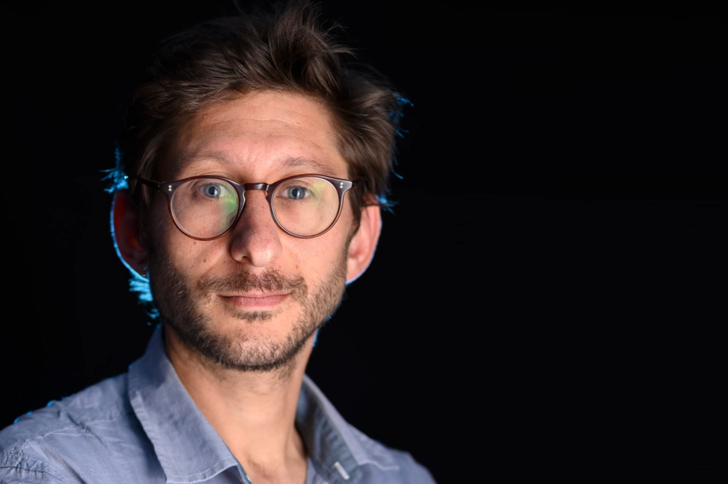 A man with light skin, short brown hair, glasses, and a beard is wearing a blue collared shirt. Against a black background, he looks at the camera with a neutral expression, supporting the call to bring Danny Fenster home.