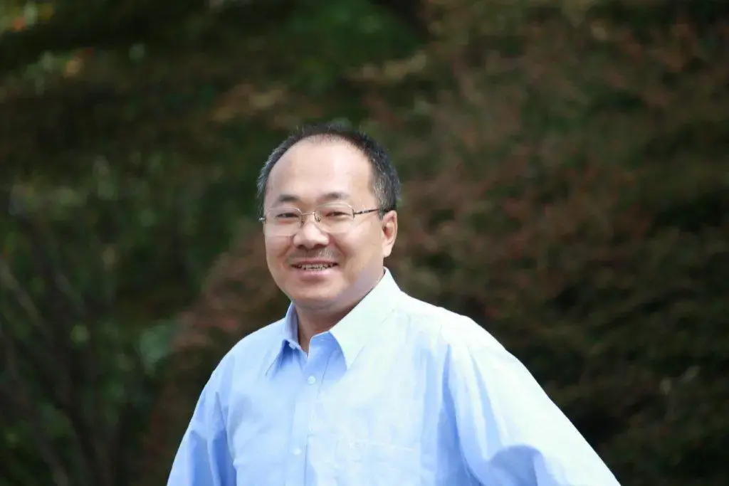 A man wearing glasses and a light blue button-up shirt smiles while standing outdoors, with green foliage blurred in the background.