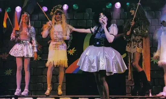Four people stand on stage in colorful, theatrical costumes under bright lights. One person in the center holds a microphone, while the others hold rainbow flags. The background is decorated with rainbow and star motifs.