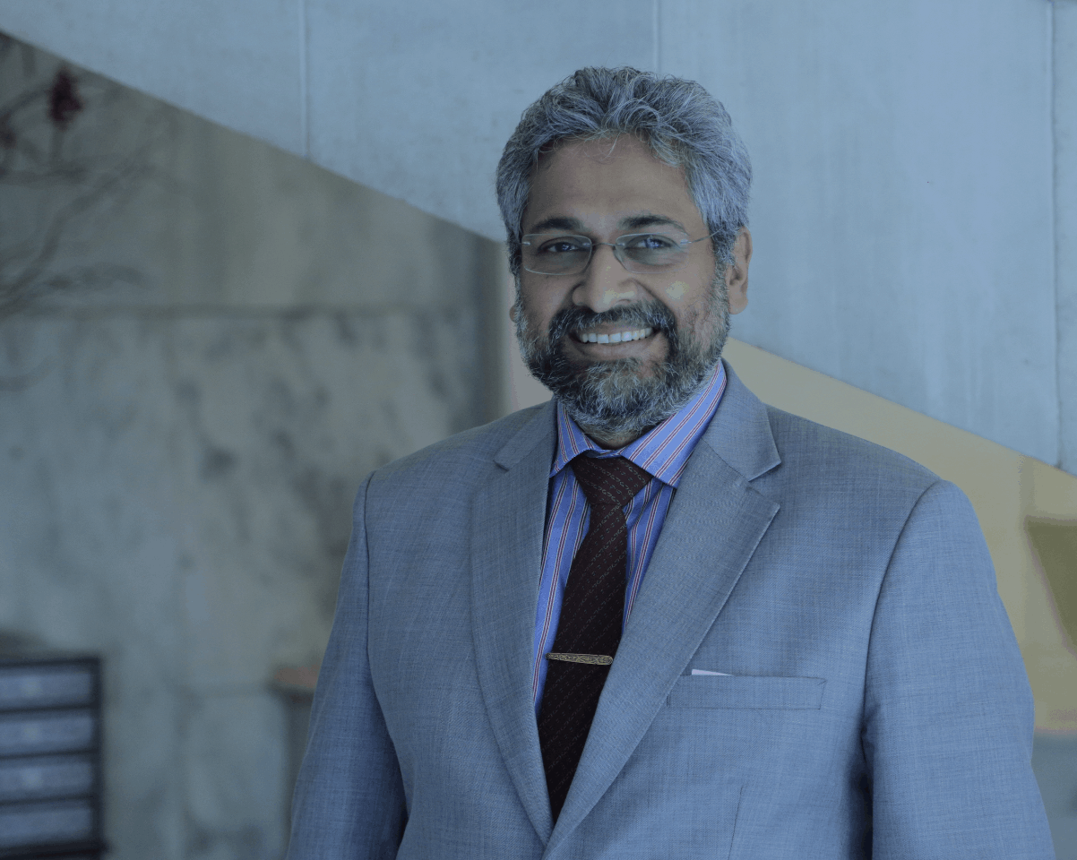 A man with gray hair and a beard, wearing glasses, a gray suit, a striped shirt, and a dark tie, smiles while standing indoors near a marble wall and staircase.