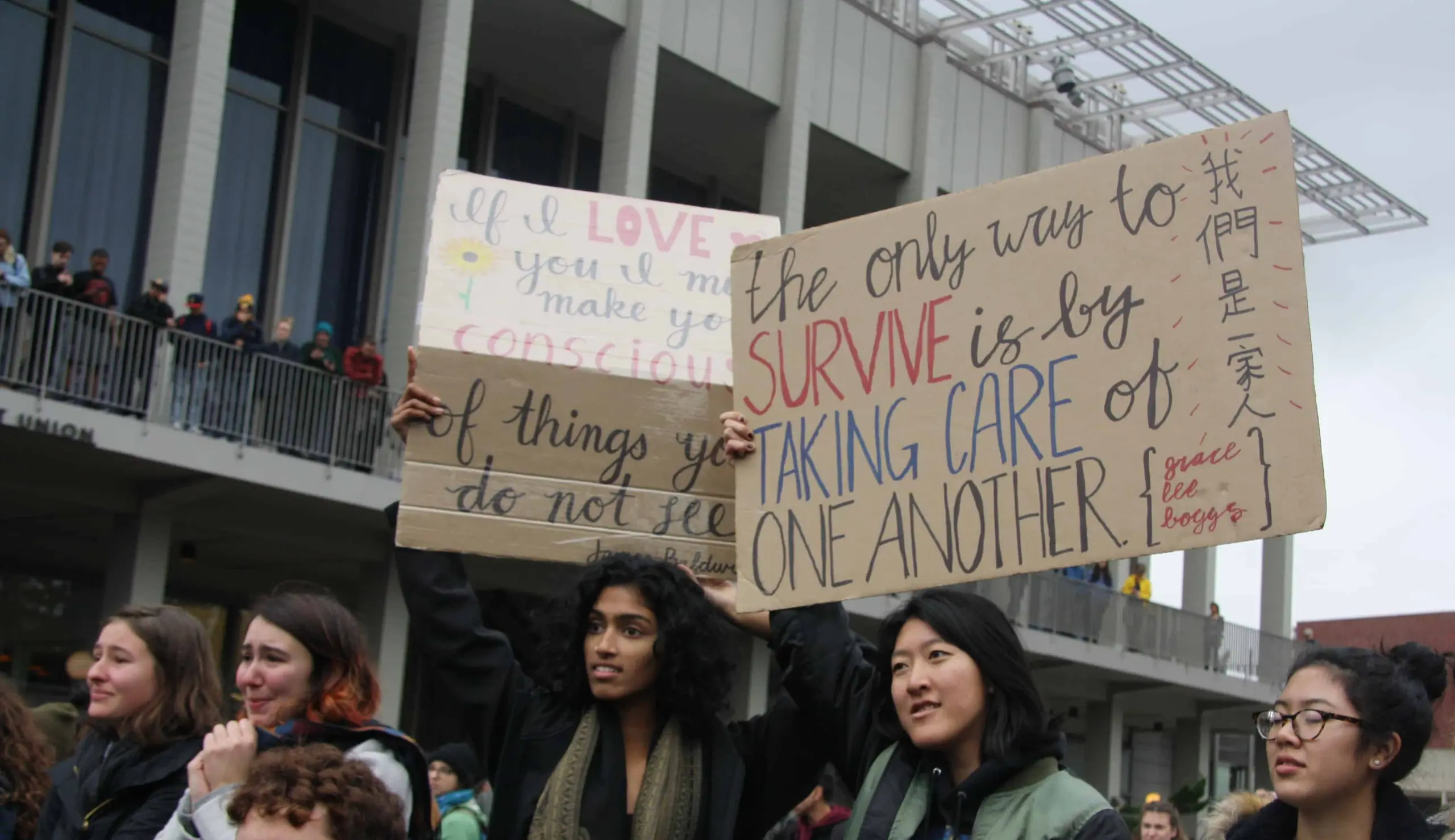 Two people in a crowd hold up protest signs about caring for one another. One sign reads, The only way to survive is by taking care of one another with some Chinese characters. Others watch from a building in the background.