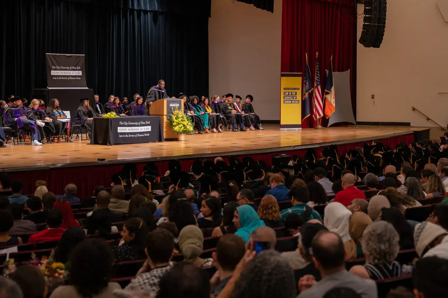 A graduation ceremony in a large auditorium shows graduates in caps and gowns seated on stage, with a speaker at a podium, and an audience of family and friends watching from the seats.