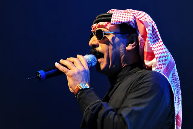 A man wearing sunglasses, a red and white keffiyeh, and a black outfit sings into a microphone on stage against a dark blue background.
