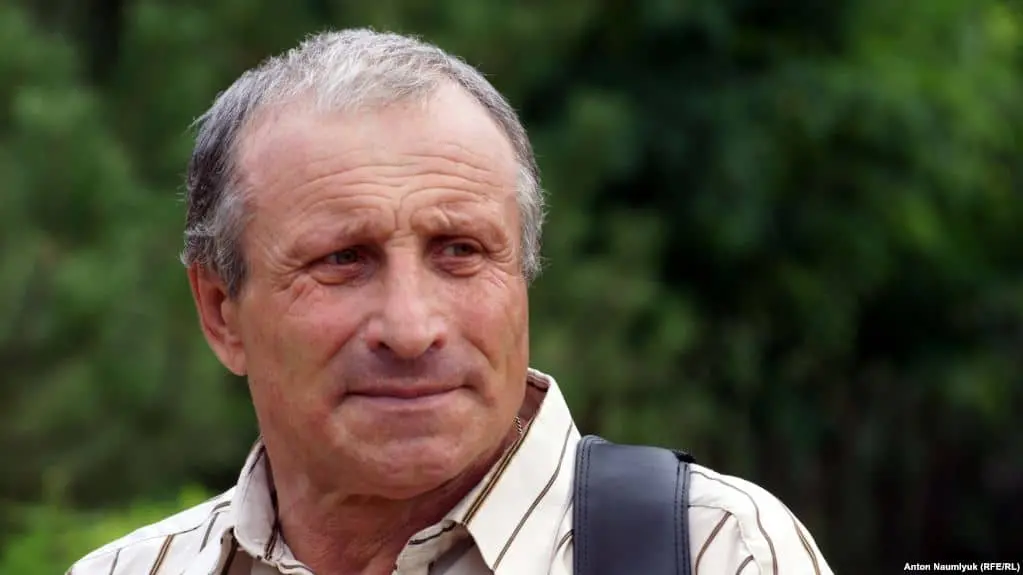 An older man with short gray hair and a striped shirt stands outdoors, looking slightly to the side with a neutral expression. The background is blurred with greenery.