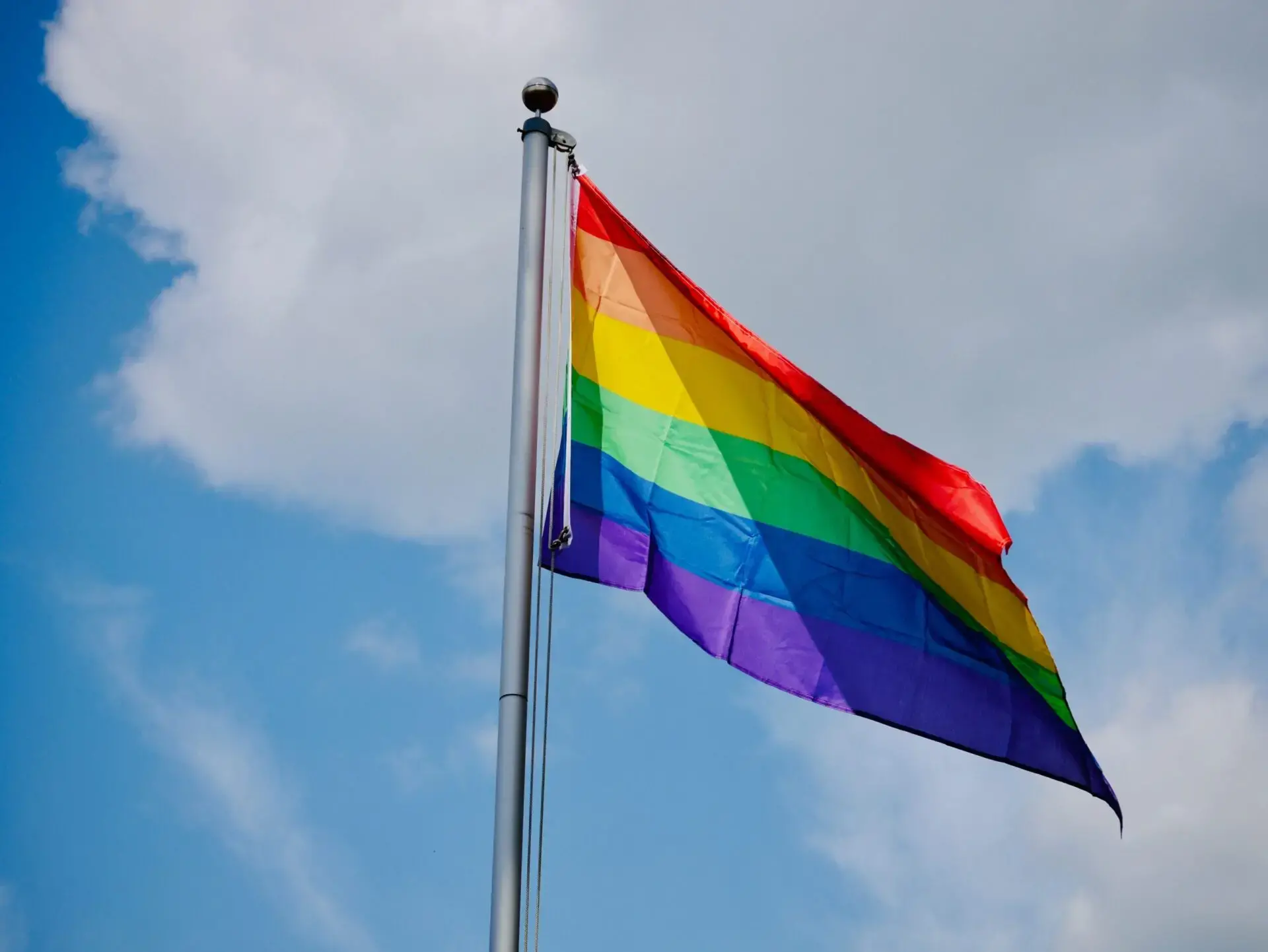 A rainbow-colored pride flag waving on a flagpole against a blue sky with clouds.
