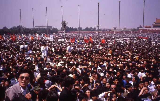 A large crowd of people fills a public square, many holding banners and flags. The scene is lively and dense, with tall flagpoles in the background and historic buildings visible in the distance.