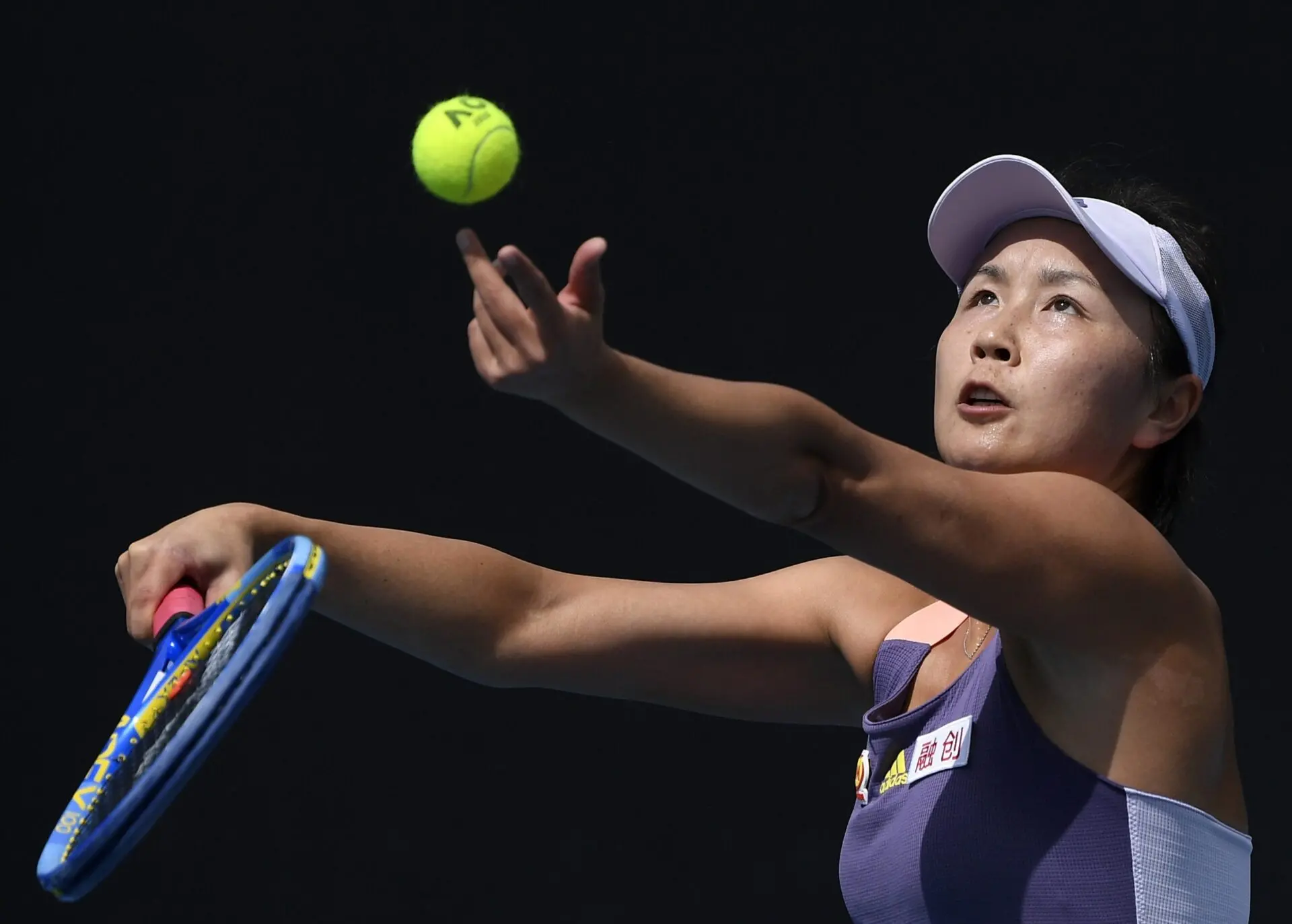 A female tennis player in a purple outfit tosses a tennis ball into the air, preparing to serve during a match, with a focused expression on her face.