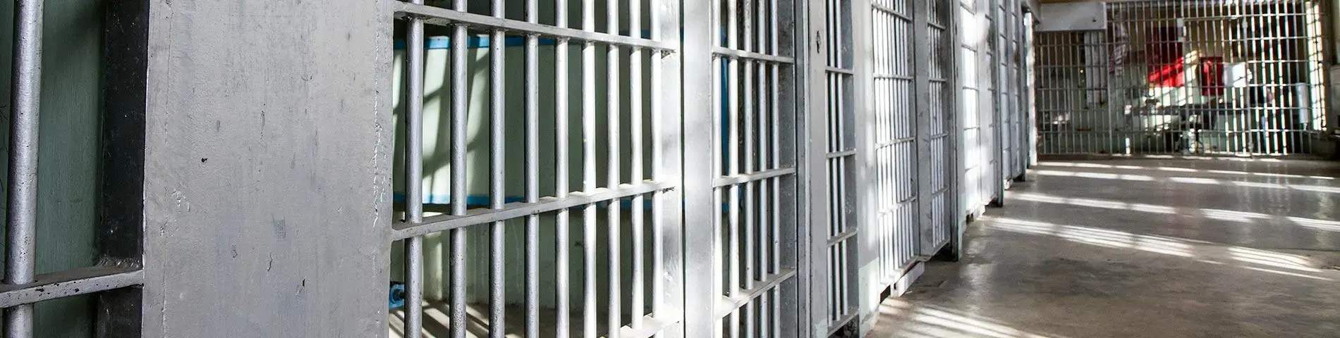 Rows of empty jail cells with metal bars line both sides of a corridor. Sunlight streams through windows, casting shadows on the shiny floor. The area appears clean but stark and institutional.