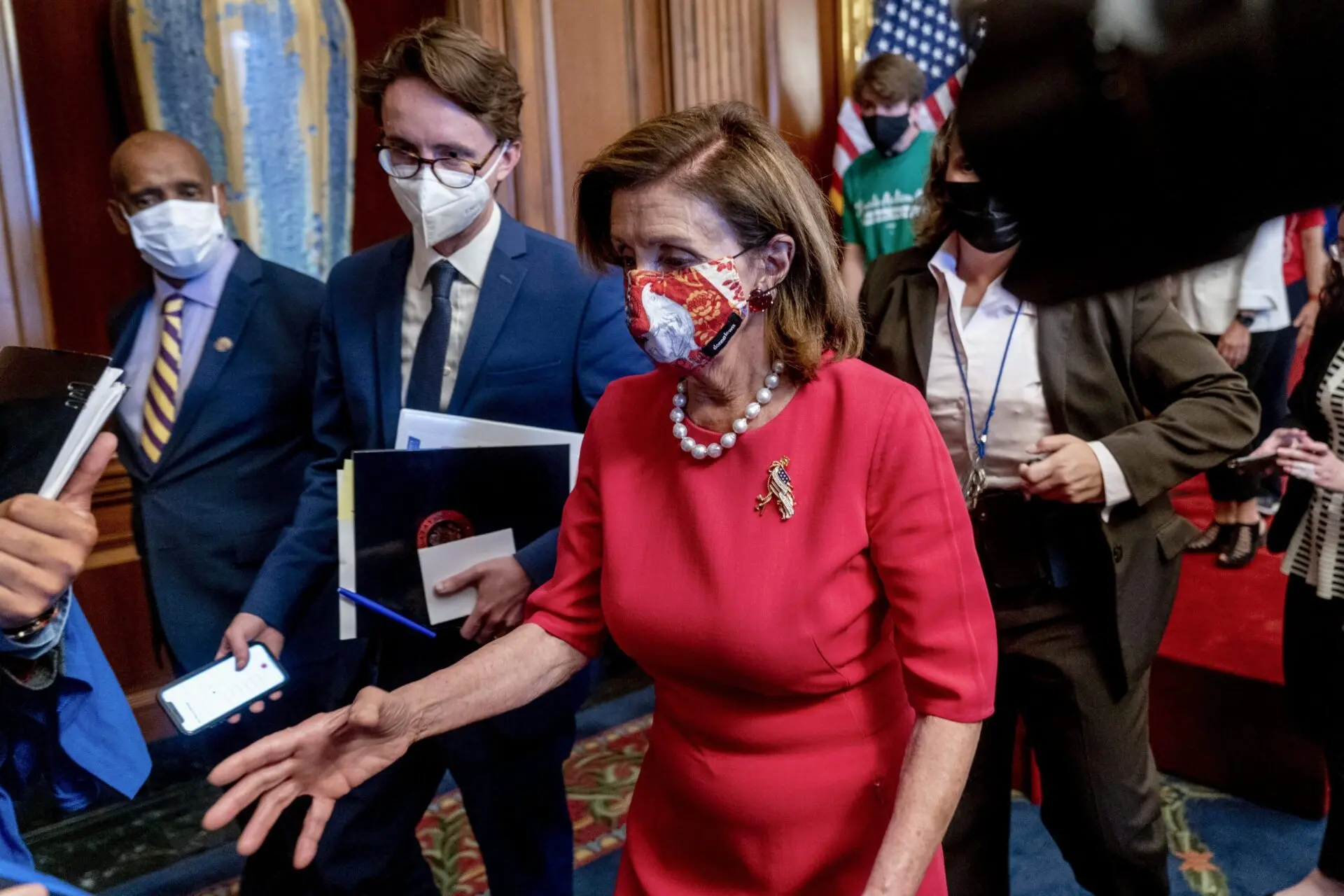 A woman in a red dress and patterned face mask walks through a group of people, some wearing masks and holding documents, in a formal room with U.S. flags and ornate decorations in the background.