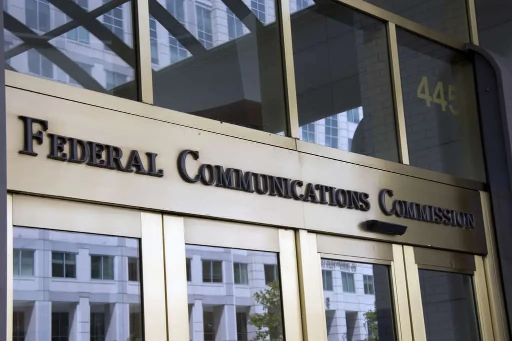 Entrance to a building with a glass door displaying the words Federal Communications Commission in bold letters, with a reflection of surrounding buildings visible in the glass.