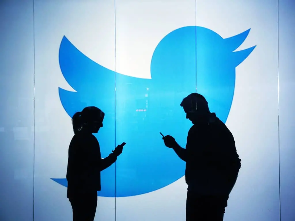 Two people stand facing each other, both using their phones, silhouetted in front of a large blue Twitter bird logo on a white background.