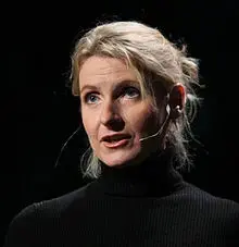 A woman with blonde hair in a loose updo wears a black turtleneck and a microphone headset, speaking against a dark background.