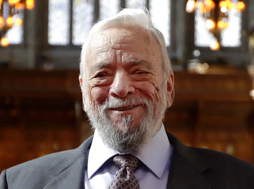 An older man with white hair and a beard, wearing a suit and tie, smiling warmly indoors with soft, warm lighting and stained glass windows in the background.
