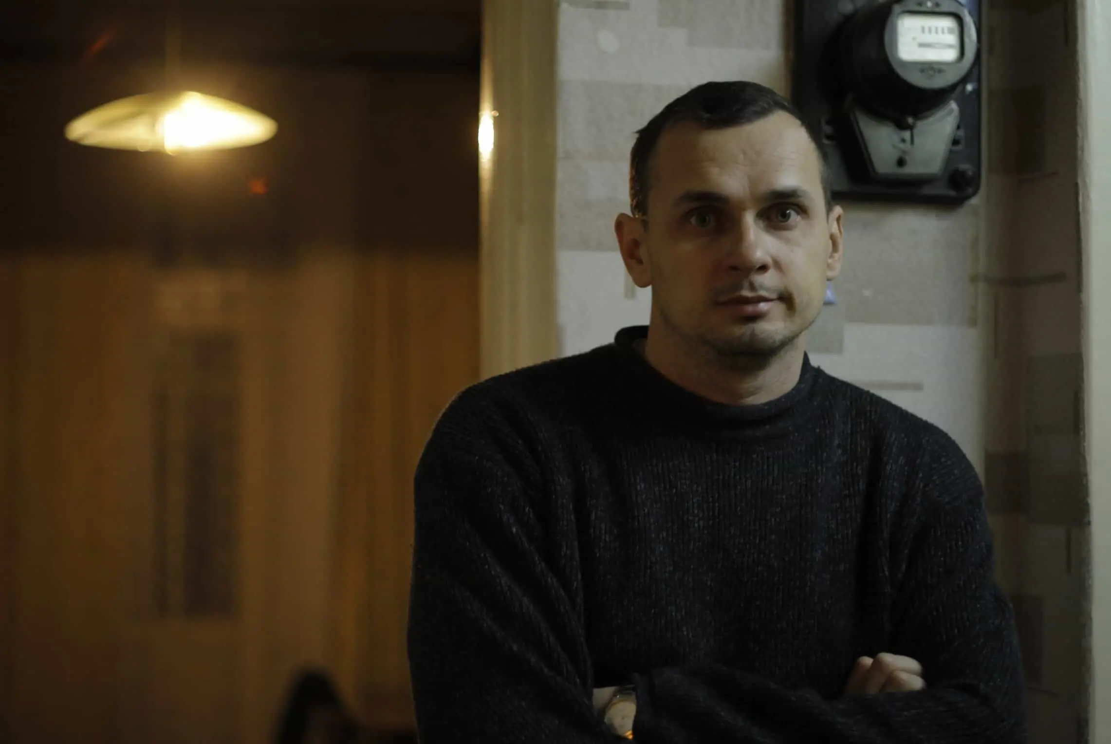 A man with short dark hair, wearing a dark sweater, stands indoors with his arms crossed. He looks directly at the camera. A round electric meter is mounted on the wall behind him. Warm light fills the background.