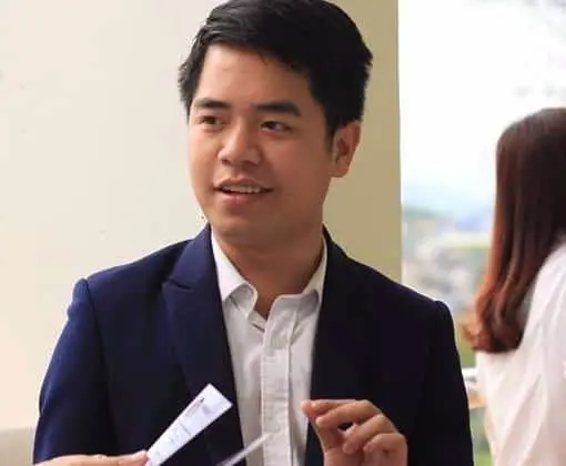 A man in a dark blue suit and white shirt is seated indoors, holding a small paper or card, and speaking. Another person is blurred in the background.