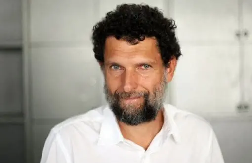 A man with curly dark hair, a beard, and blue eyes is smiling slightly. He is wearing a white collared shirt and is posed in front of a neutral, light-colored background.