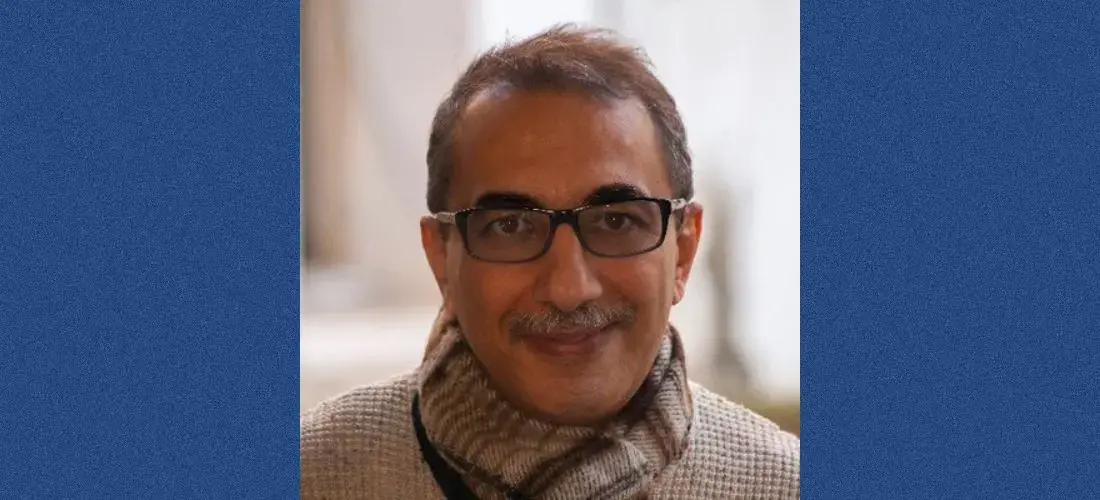 A middle-aged man with short hair, wearing glasses and a beige scarf, smiles at the camera. The background is softly blurred, and the image is bordered by blue vertical bars on each side.
