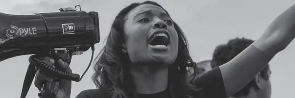 A woman holding a megaphone shouts passionately, raising one arm in the air during what appears to be a protest or rally. The image is in black and white.