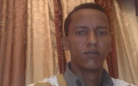 A man with short dark hair and a neutral expression looks at the camera. He is wearing a light-colored garment over a collared shirt, and there are brown and beige curtains in the background.