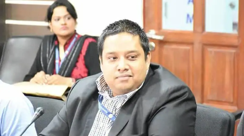 A man in a suit sits at a conference table, looking slightly to the side. A woman in professional attire sits in the background, also at the table. Both appear to be participating in a meeting or discussion.