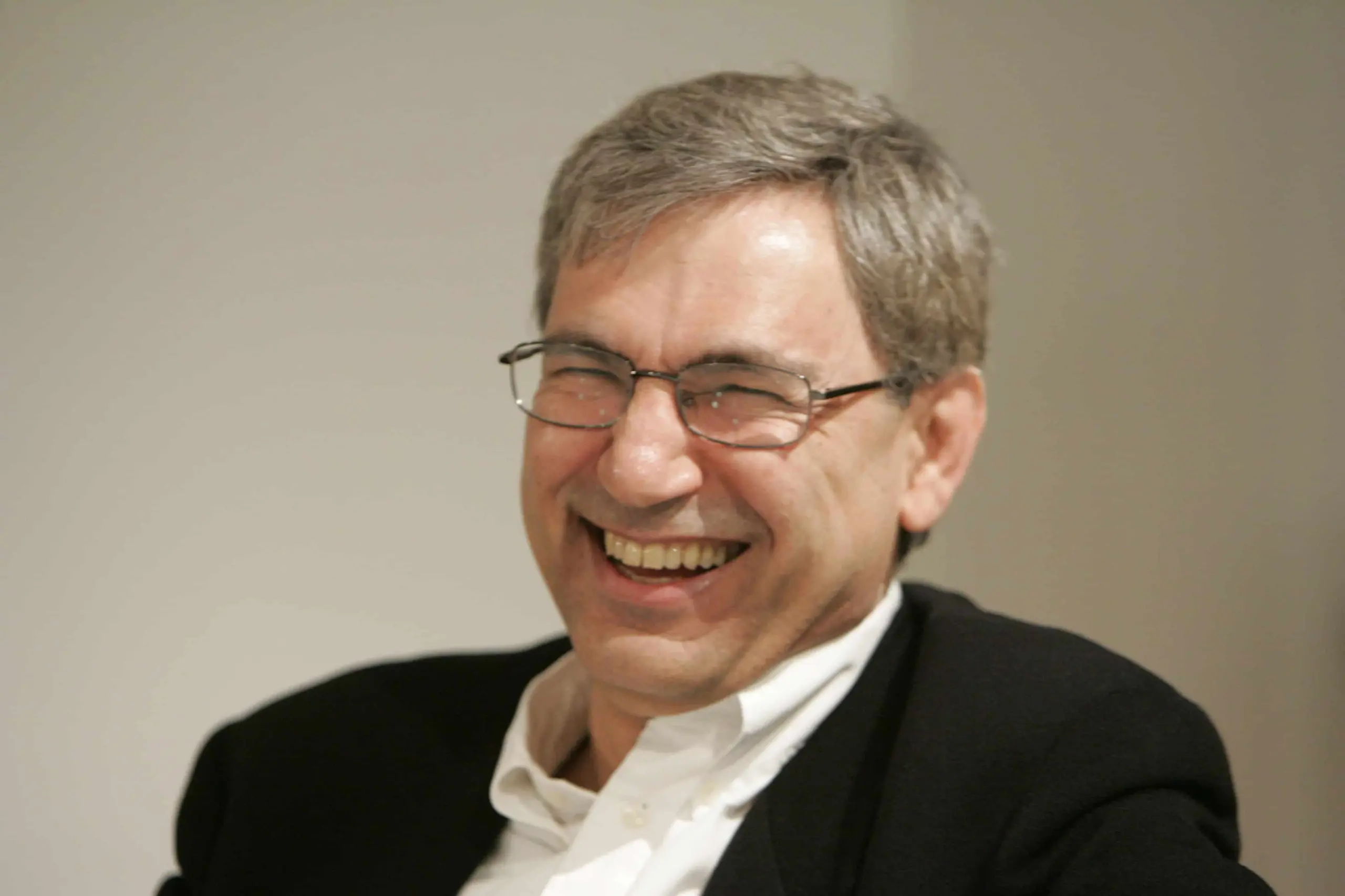A middle-aged man with short gray hair and glasses smiles broadly while wearing a black jacket and white shirt against a neutral background.