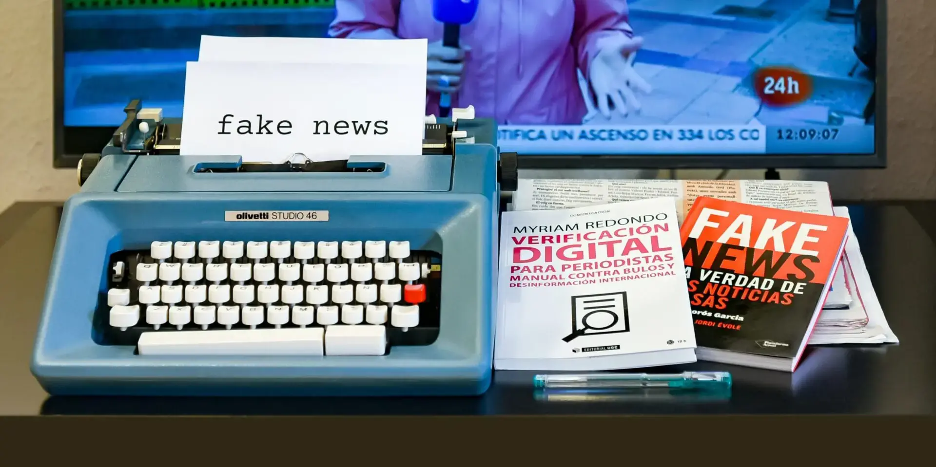 A blue typewriter with a paper reading fake news sits on a table next to books about digital verification and fake news, with a TV showing a news broadcast in the background.