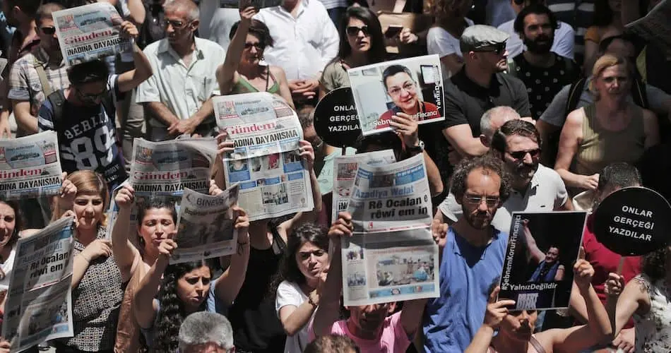 A large group of people protest outdoors, holding up newspapers and posters with photos and text. Many wear sunglasses, and most look serious or determined. The crowd is closely packed under daylight.